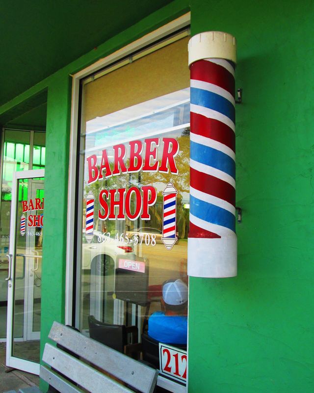 Bir berber dükkânının cephesi: camında BARBER SHOP yazısı, kapının yanında boyalı bir berber direği.
